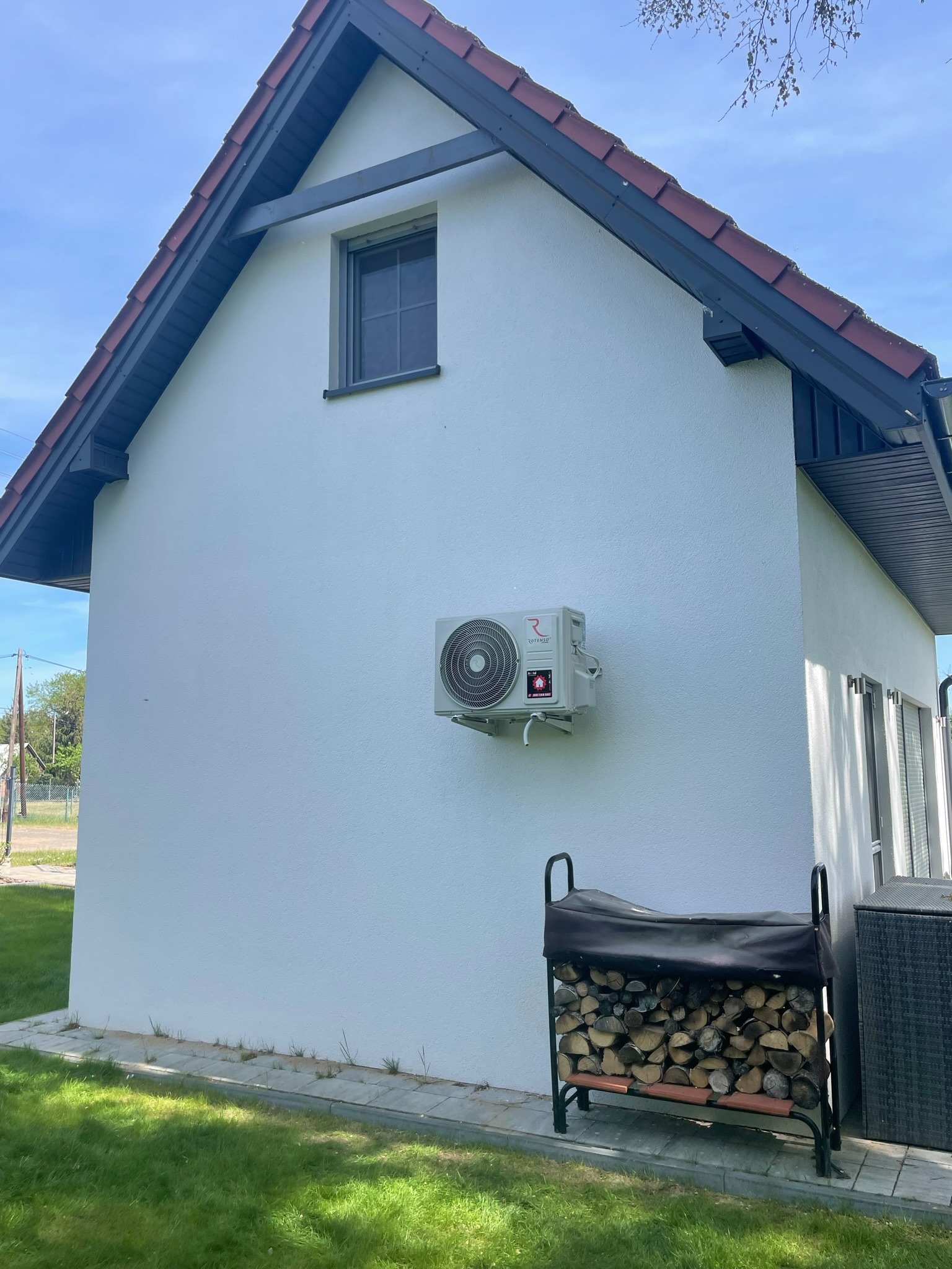 Biały budynek z zamontowaną na ścianie jednostką zewnętrzną klimatyzacji marki Rotenso, widoczny skład drewna na trawniku.