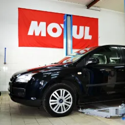 Czarny samochód na podnośniku w warsztacie, widoczny baner reklamowy Motul na ścianie, w tle wózek narzędziowy i butle.