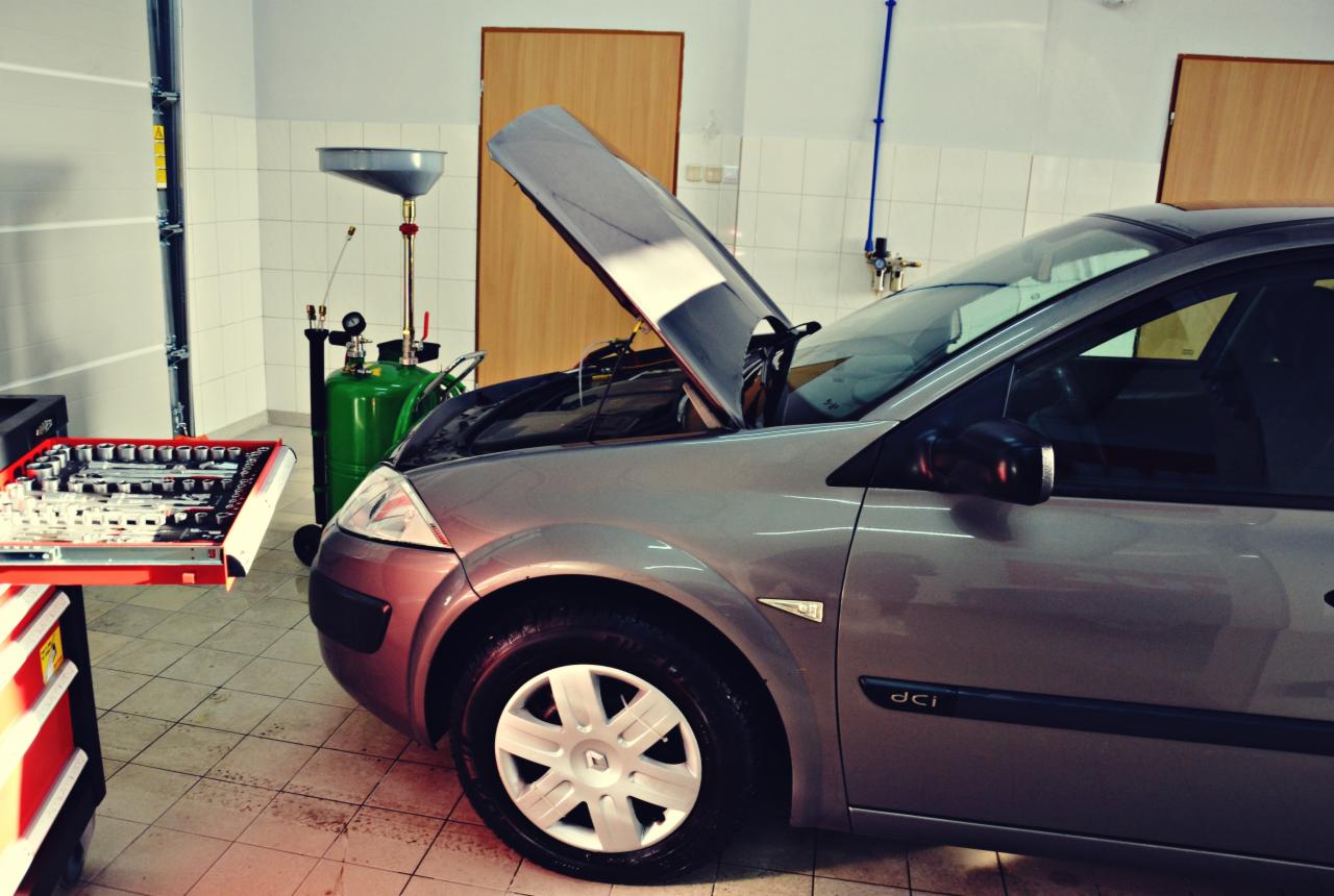 Szary samochód z otwartą maską w warsztacie, widoczny wózek z narzędziami, urządzenie do wymiany oleju i ceramiczna podłoga