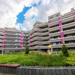 Nowoczesny budynek apartamentowy z balkonami w odcieniach szarości i różu, z zielonym dziedzińcem, placem zabaw i ścieżką spacerową.