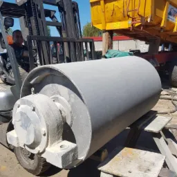 Szary, cylindryczny element maszyny z łożyskiem, podnoszony przez wózek widłowy na zewnątrz, widoczne ślady korozji i przygotowania do obróbki powierzchni.