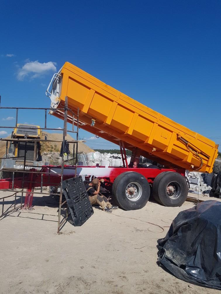 Żółta przyczepa wywrotka w trakcie malowania, z widocznym pracownikiem nanoszącym farbę na czerwoną ramę, na tle błękitnego nieba z chmurami.