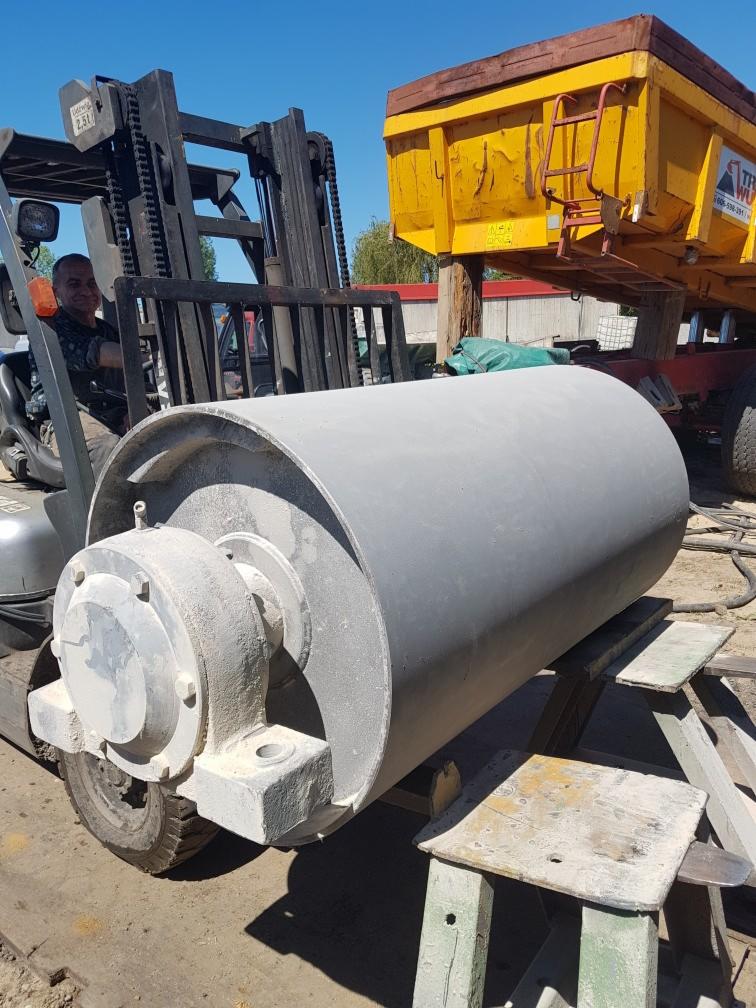 Szary, cylindryczny element maszyny z łożyskiem, podnoszony przez wózek widłowy na zewnątrz, widoczne ślady korozji i przygotowania do obróbki powierzchni.