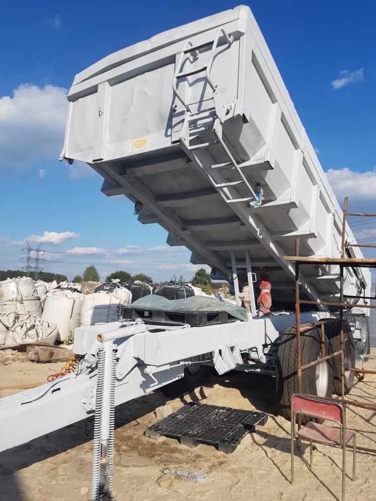 Wywrócona naczepa ciężarówki po odświeżeniu powłoki, widoczne drabinki i elementy konstrukcyjne, w tle worki z materiałem i niebieskie niebo.