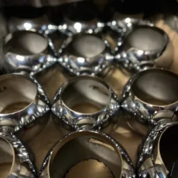 Partia chromowanych pierścieni metalowych, prawdopodobnie tulei lub elementów maszyn, ułożonych w kartonowym pudełku, widoczne refleksy światła na polerowanej powierzchni.