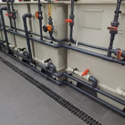Szare rury przemysłowe z pomarańczowymi zaworami i przepływomierzami, zamontowane wzdłuż ściany nad podłogą z odpływem liniowym.