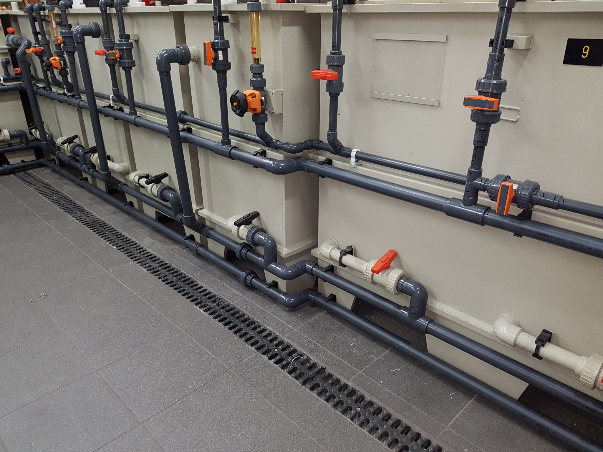 Szare rury przemysłowe z pomarańczowymi zaworami i przepływomierzami, zamontowane wzdłuż ściany nad podłogą z odpływem liniowym.