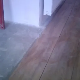 Połączenie nowej drewnopodobnej podłogi z surowym betonem przy ścianie z grzejnikiem; widoczny czerwony profil wykończeniowy.