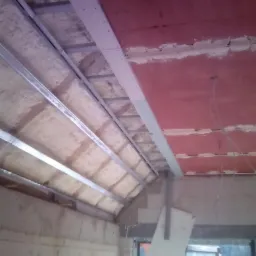 Wnętrze w trakcie prac: metalowy stelaż pod płytę gipsowo-kartonową na suficie, widoczna izolacja piankowa, fragment ściany z czerwonym podkładem i instalacją elektryczną.