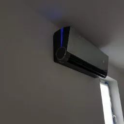 Czarny klimatyzator ścienny z niebieskim podświetleniem zamontowany w pomieszczeniu, widoczne okno z zabezpieczeniami malarskimi.