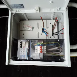 Otwarta skrzynka elektryczna z widocznymi bezpiecznikami, przewodami i elementami montażowymi. W tle widoczne rury PCV.