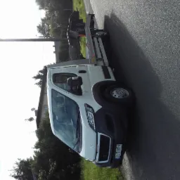 Biała laweta na drodze asfaltowej, częściowo zacieniona, z platformą załadunkową i widoczną tablicą rejestracyjną EL 5A834.