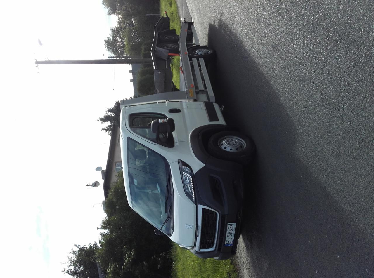 Biała laweta na drodze asfaltowej, częściowo zacieniona, z platformą załadunkową i widoczną tablicą rejestracyjną EL 5A834.