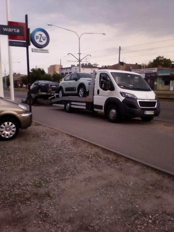 Laweta z dwoma autami na tle miejskiego krajobrazu w Pabianicach. Biały bus z tablicą rejestracyjną BT 131SN. Słup Warta i PZU Pomoc.