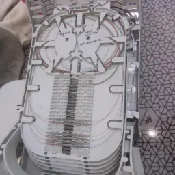 Otwarta skrzynka z kablami światłowodowymi, widoczne delikatne kolorowe włókna ułożone wewnątrz białej plastikowej obudowy, obok leżą robocze rękawice.