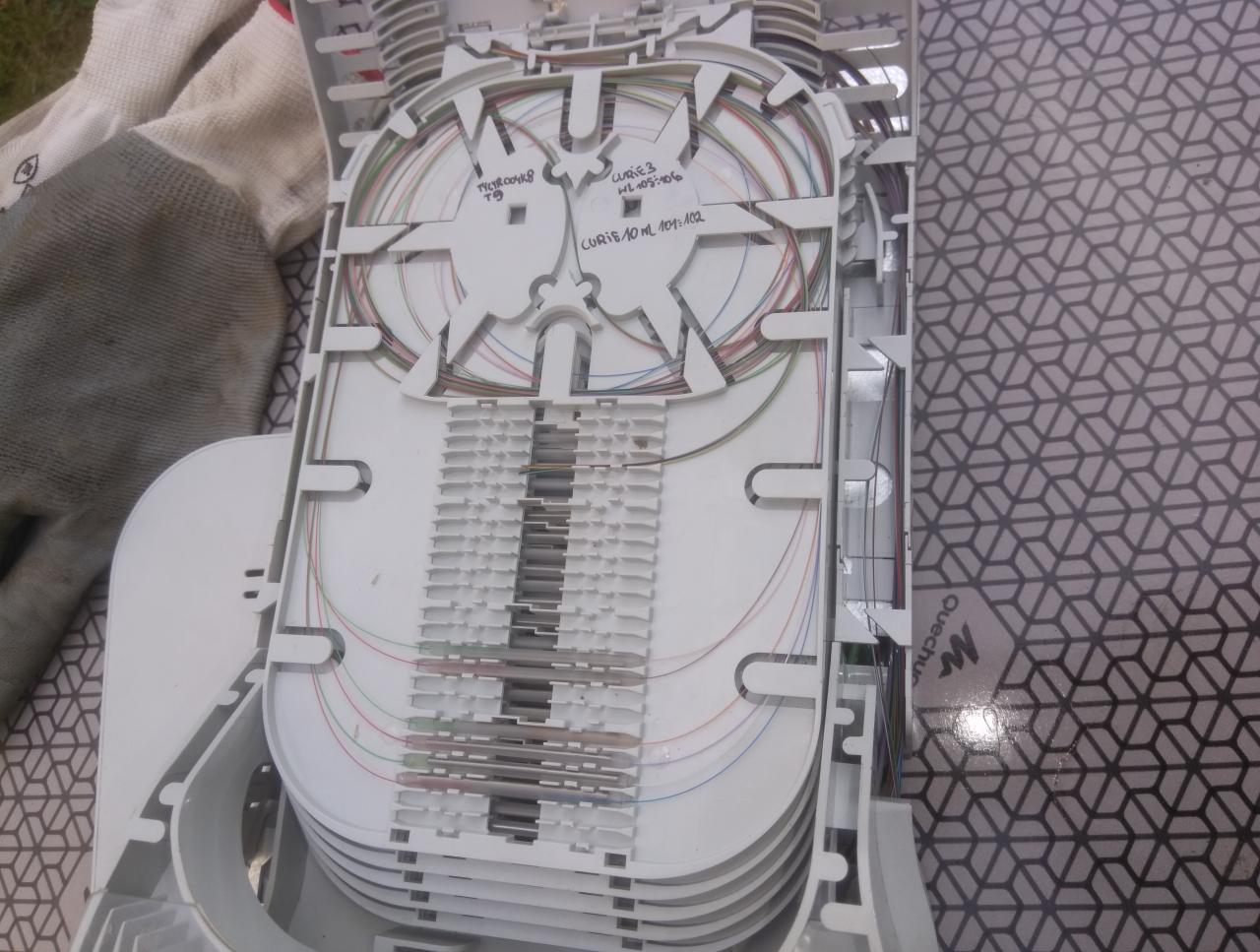 Otwarta skrzynka z kablami światłowodowymi, widoczne delikatne kolorowe włókna ułożone wewnątrz białej plastikowej obudowy, obok leżą robocze rękawice.