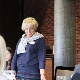 Kobieta w szarym szalu i granatowej bluzce, stojąca przy stole z nakryciami, podczas spotkania w pomieszczeniu z ceglaną ścianą i betonowym filarem.