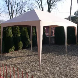 Beżowy namiot ogrodowy z metalową konstrukcją, ustawiony na żwirowym podłożu, otoczony tujami i niskim, czerwonym płotkiem. Widok z niskiej perspektywy.