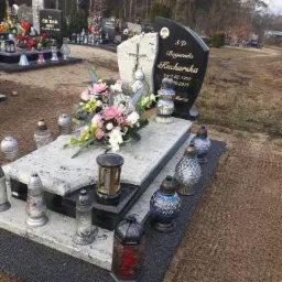 Granitowy pomnik z czarną tablicą, ozdobiony kwiatami i lampionami, na cmentarzu w pochmurny dzień.