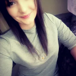 Uśmiechnięta młoda kobieta z ciemnymi włosami i naszyjnikiem z okrągłym medalionem, ubrana w szarą sukienkę, robi selfie w pomieszczeniu.