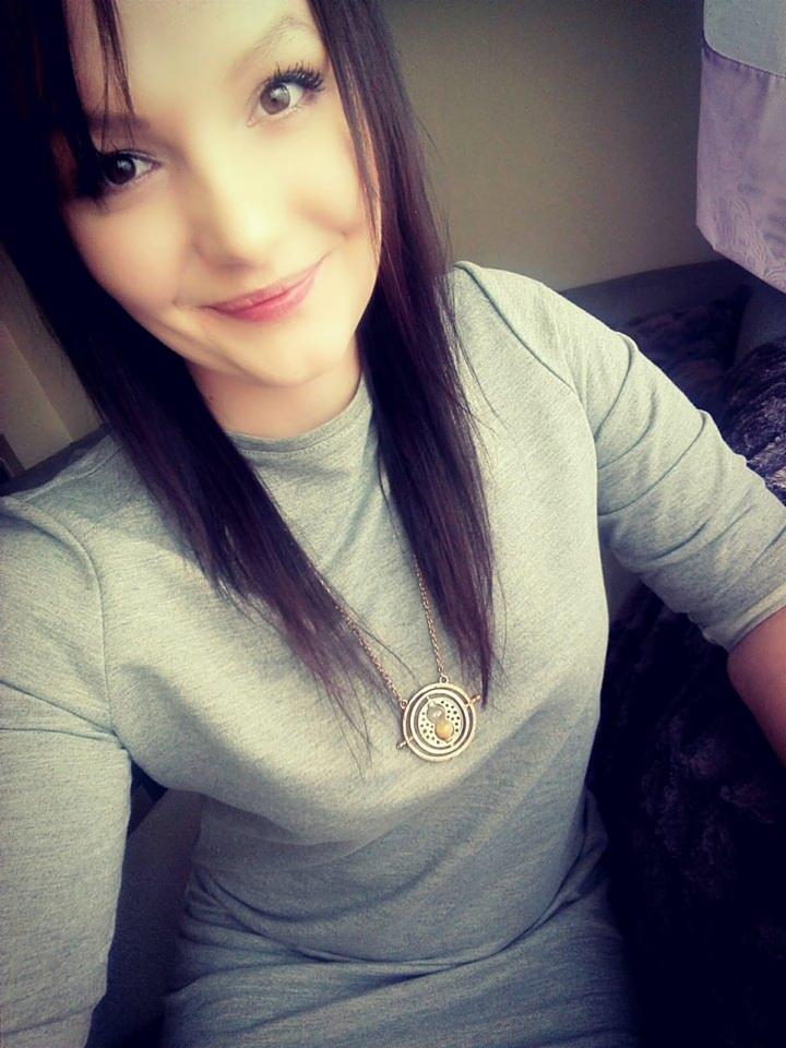 Uśmiechnięta młoda kobieta z ciemnymi włosami i naszyjnikiem z okrągłym medalionem, ubrana w szarą sukienkę, robi selfie w pomieszczeniu.