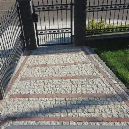 Ścieżka z jasnej kostki granitowej z trzema pasami z czerwonej kostki, prowadząca do furtki z czarnego metalu, na tle zielonego trawnika i drzew w oddali.