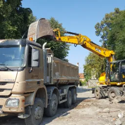 Żółta koparka JCB ładuje ziemię do wywrotki Mercedes Actros w kolorze piaskowym na tle zielonych drzew.
