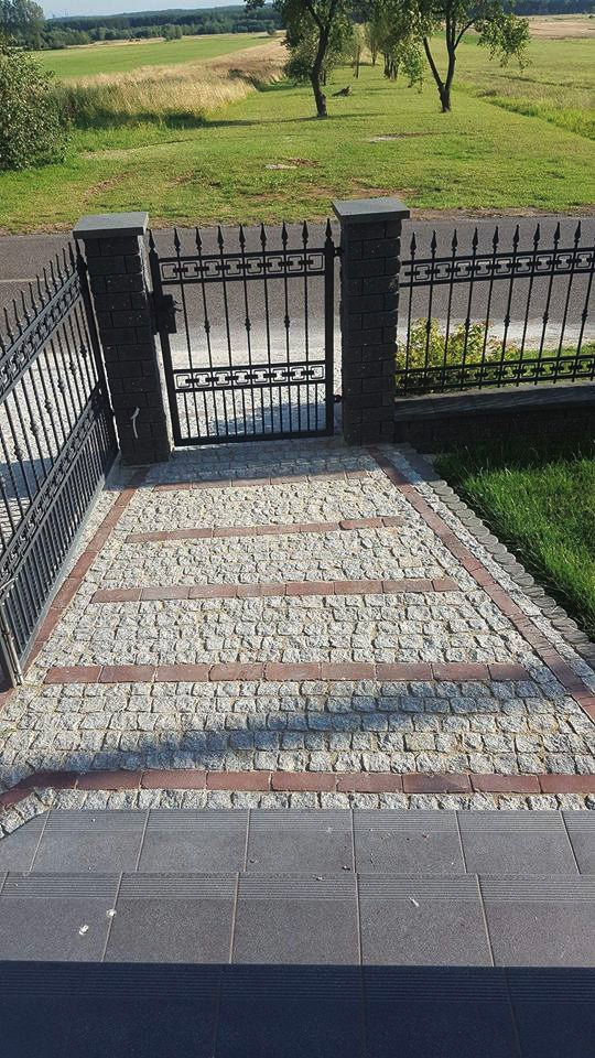Ścieżka z jasnej kostki granitowej z trzema pasami z czerwonej kostki, prowadząca do furtki z czarnego metalu, na tle zielonego trawnika i drzew w oddali.