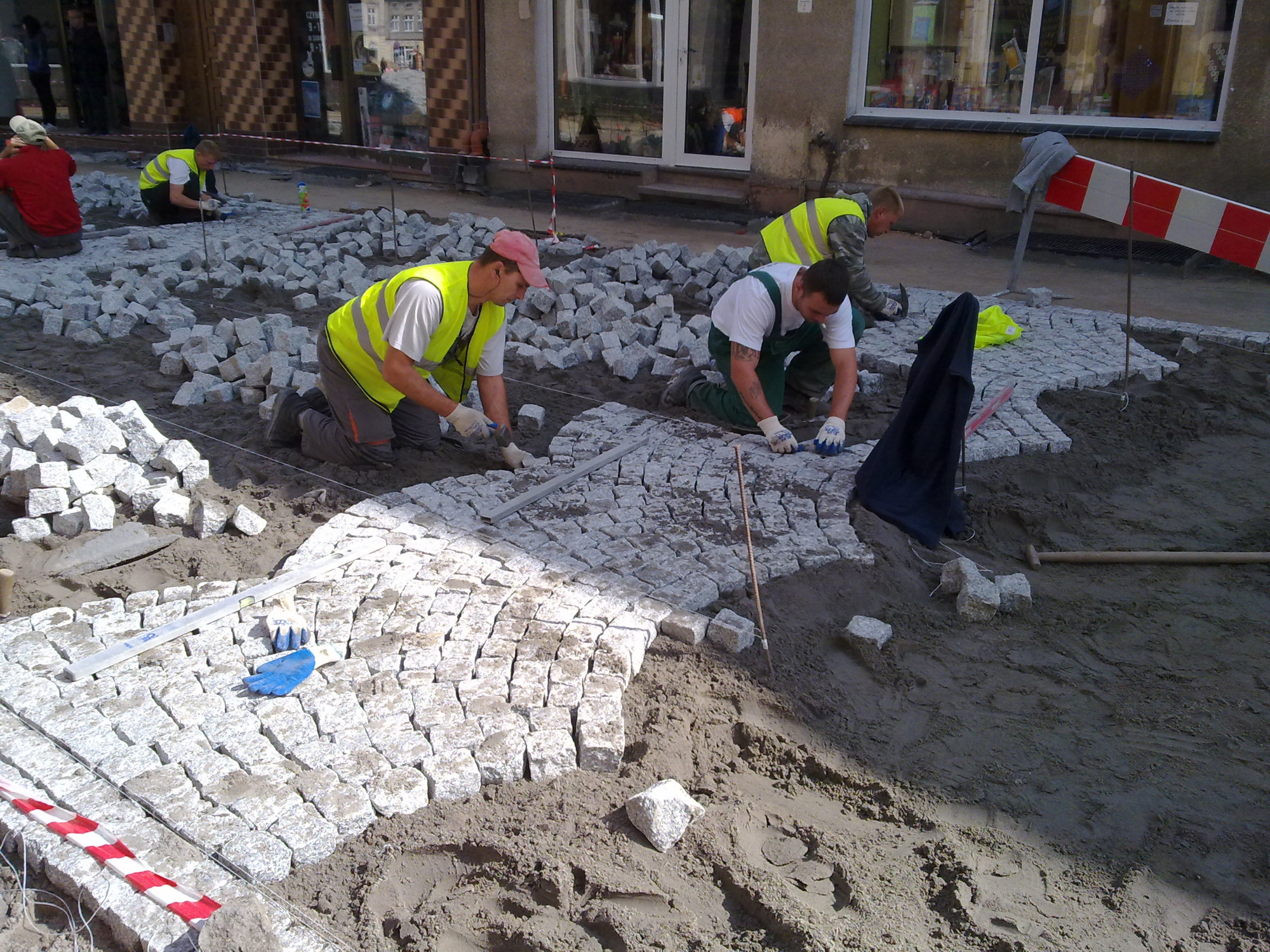 Robotnicy w kamizelkach odblaskowych układają szarą kostkę brukową na piasku, widoczny poziomica i rękawice robocze.