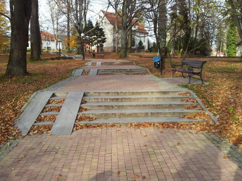 Parkowa ścieżka z kostki brukowej przechodząca w schody z rampami dla wózków, otoczona drzewami i opadłymi liśćmi.