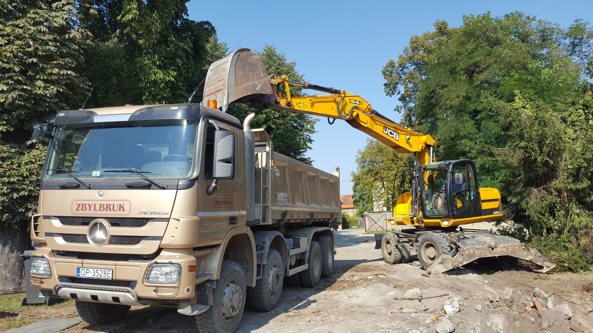 Żółta koparka JCB ładuje ziemię do wywrotki Mercedes Actros w kolorze piaskowym na tle zielonych drzew.