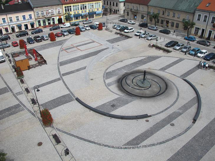Widok z góry na rynek miejski z fontanną w kształcie spirali, wyłożony kostką brukową w geometryczny wzór, otoczony budynkami i zaparkowanymi samochodami.