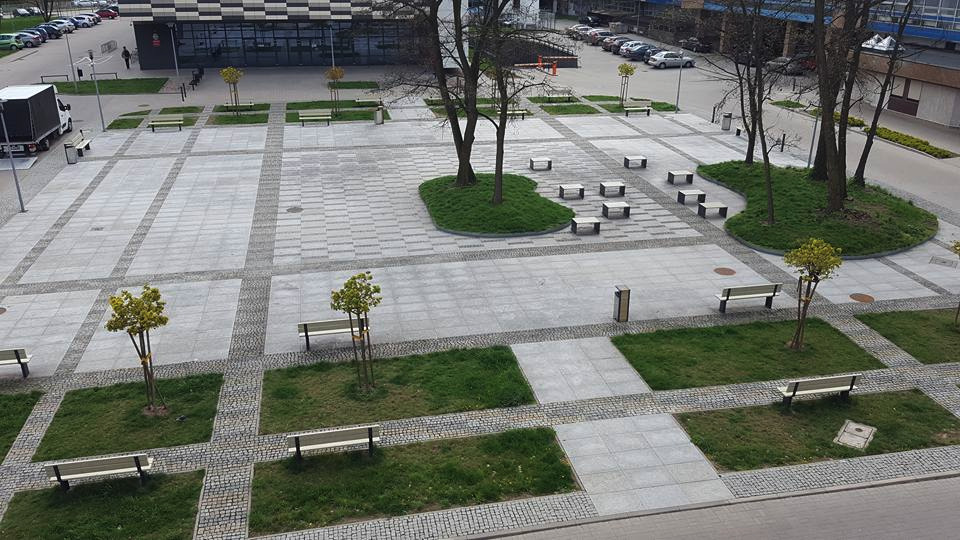 Rozległy plac wyłożony jasnoszarą kostką brukową w geometryczny wzór, z kwadratowymi trawnikami, ławkami i młodymi drzewami, widziany z góry.