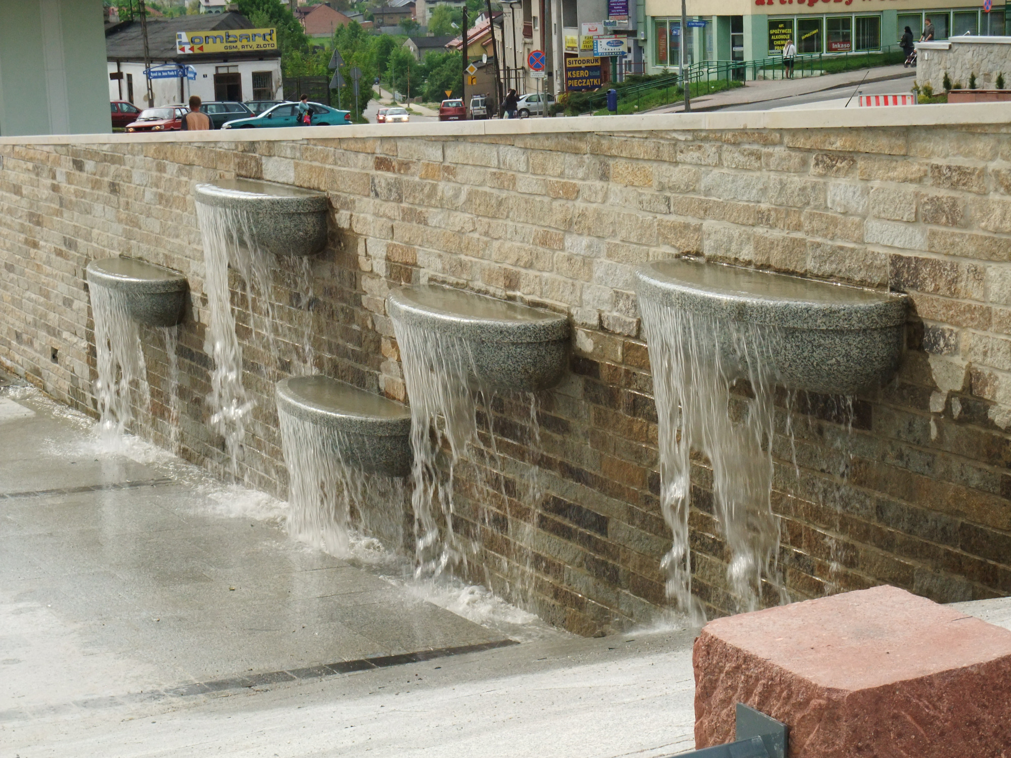 Kaskadowa fontanna miejska z kamiennymi misami na murowanej ścianie, w tle zabudowa miejska i zieleń.