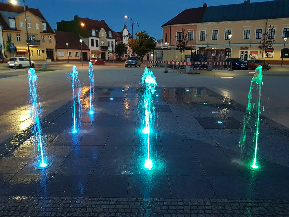 Podświetlana fontanna posadzkowa z dyszami na placu miejskim, widok na budynki w tle.