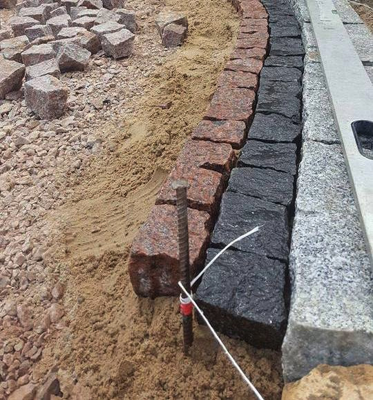 Trzy rzędy kostki brukowej w różnych kolorach (czerwony, czarny, szary) układane na piasku, wyznaczane sznurkiem i poziomnicą. W tle widoczne kamienie i gruz.