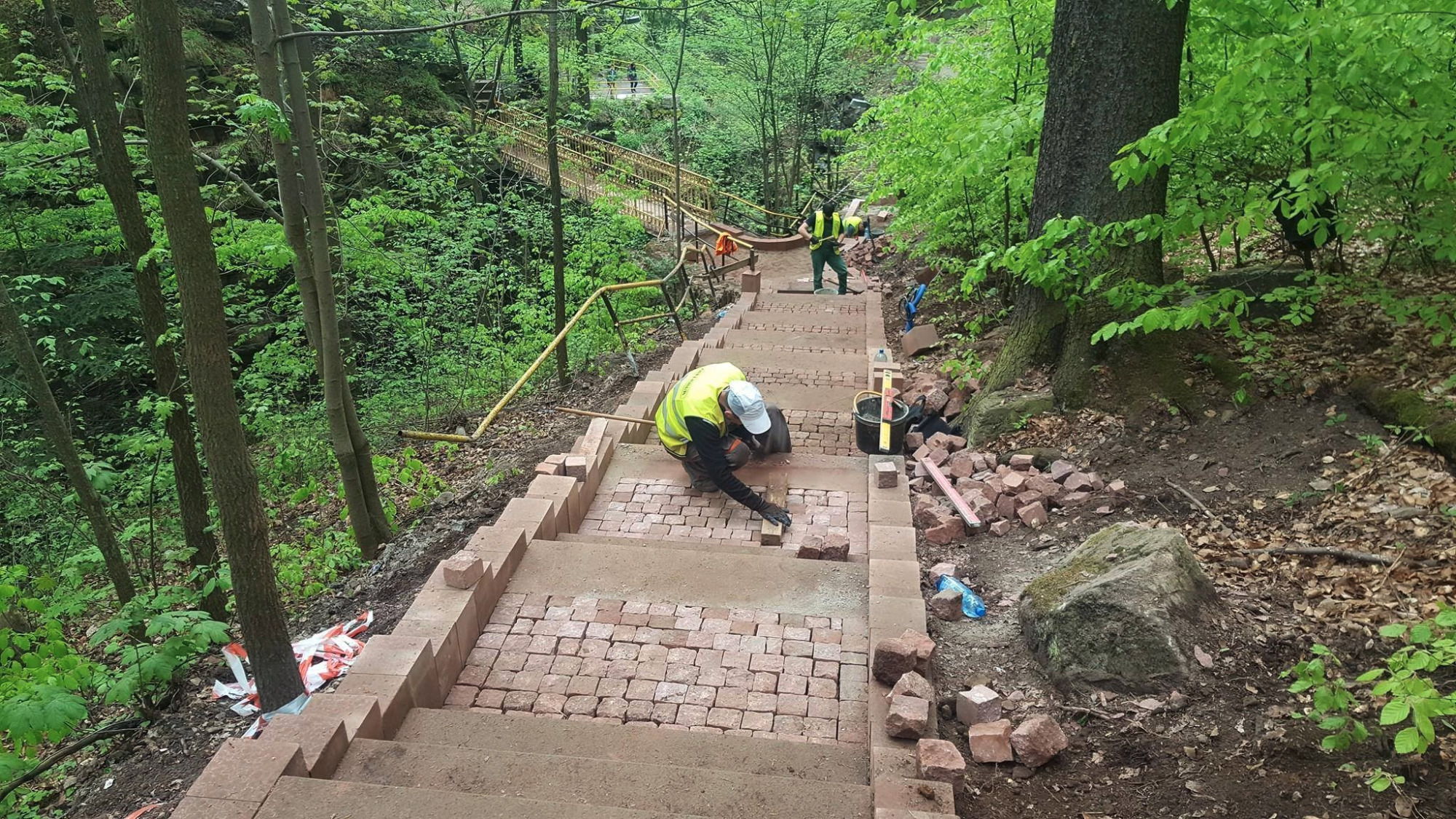 Robotnicy układają czerwoną kostkę brukową na schodach w lesie, widoczny most w tle.
