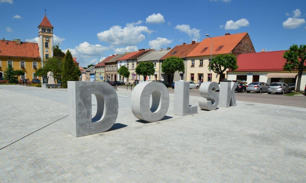 Betonowy napis 'DOLSK' na wybrukowanym placu miejskim w słoneczny dzień, z budynkami i wieżą w tle.