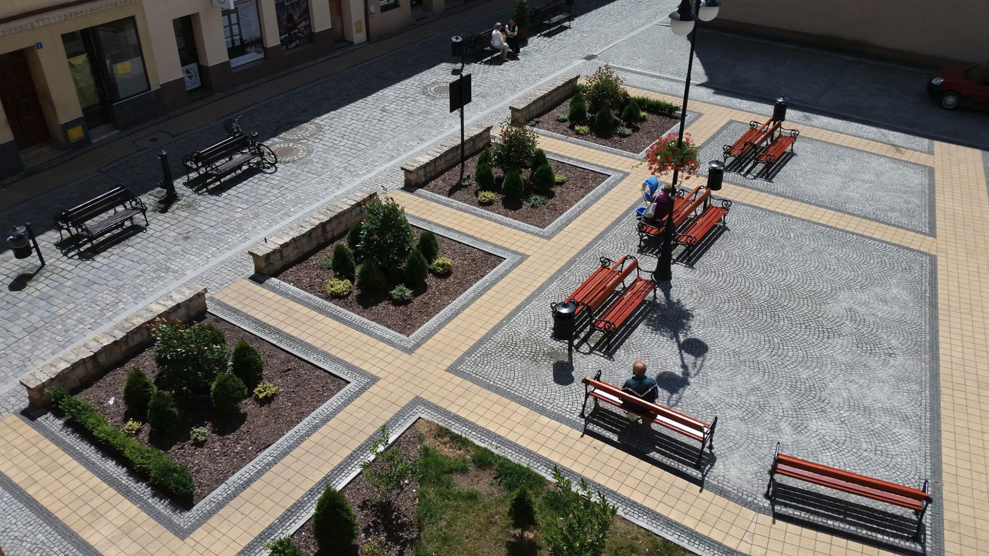Widok z góry na plac wyłożony kostką brukową w różnych kolorach, z rabatami kwiatowymi, ławkami i siedzącymi ludźmi.