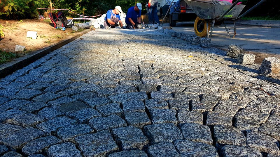 Układanie kostki granitowej na drodze dojazdowej, widoczni pracownicy w trakcie pracy, taczka z kostką w tle, częściowo ułożona nawierzchnia.