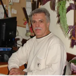 Portret biegłego w swetrze, siedzącego przy biurku z komputerem i kalendarzem w tle. Widoczne rośliny doniczkowe i dokumenty.