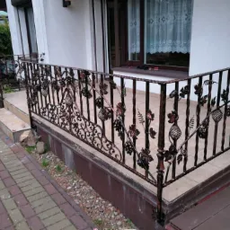 Kuta, metalowa balustrada balkonowa z motywem winogron, w kolorze miedzi, zamontowana przy wejściu do domu z jasną elewacją i kostką brukową.
