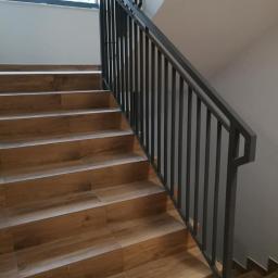 Badura Welding - Klatka schodowa z drewnopodobnymi płytkami i szarą metalową barierką z pionowymi prętami, widok z dołu schodów, w tle okno i dekoracja w czarno-białe pasy.