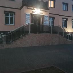 Badura Welding - Zewnętrzne schody wejściowe do budynku z balustradami ze stali nierdzewnej i szkła, oświetlenie schodów punktowe, widok na elewację budynku w odcieniach beżu i brązu.