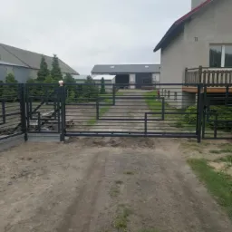 Nowoczesna, metalowa brama wjazdowa z geometrycznym wzorem poziomych prętów, zamontowana na betonowej podmurówce, prowadząca na żwirowy podjazd przed domem z drewnianym balkonem.