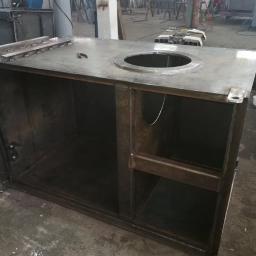 Badura Welding - Metalowa konstrukcja szafki z blachy z okrągłym otworem na blacie, widoczne spawy i surowy stan wykończenia, w tle warsztat z butlą spawalniczą i pracującymi osobami.