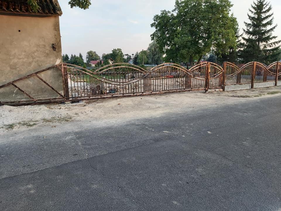 Fragment rustykalnego ogrodzenia z metalowymi prętami o falistej górnej krawędzi, częściowo opartego o budynek, z widoczną drogą i zielenią w tle.