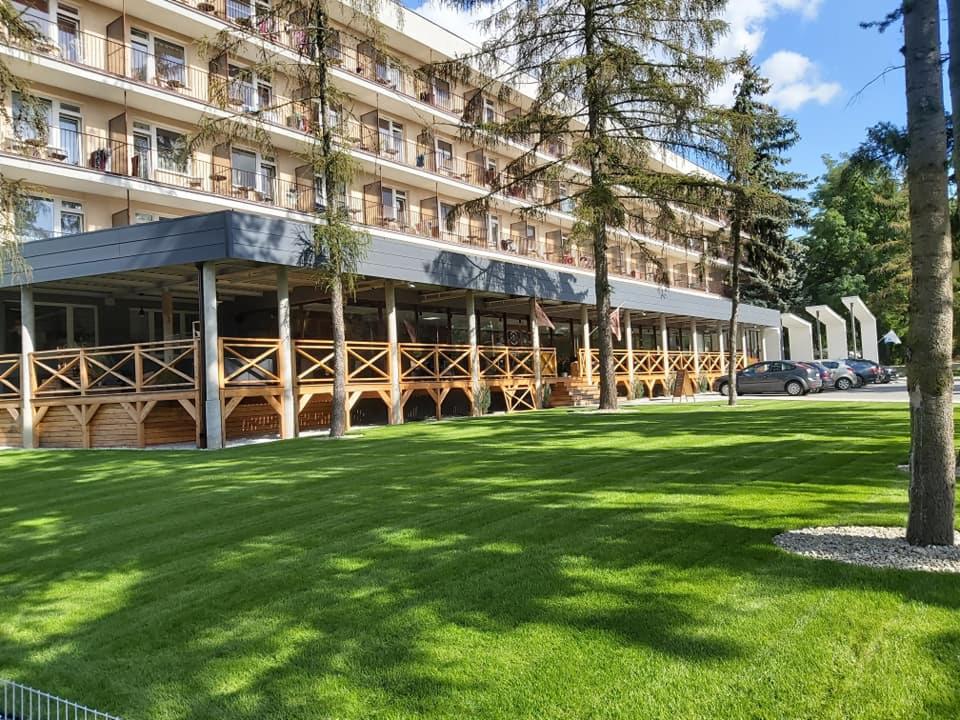 Elewacja hotelu z balkonami i drewnianą konstrukcją tarasu na parterze, widok z zadbanego trawnika w słoneczny dzień.