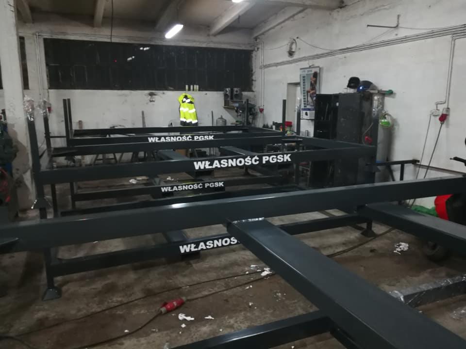 Konstrukcja metalowa z napisem 'WŁASNOŚĆ PGSK' na hali produkcyjnej, widoczny pracownik w kamizelce odblaskowej w tle.