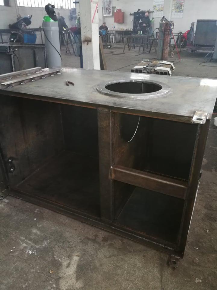 Metalowa konstrukcja szafki z blachy z okrągłym otworem na blacie, widoczne spawy i surowy stan wykończenia, w tle warsztat z butlą spawalniczą i pracującymi osobami.